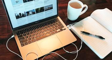 Horizontal photo of a laptop open on a wood grain desk, staged with headphone cords, an open notebook, pen, and mug. Original photo credit Alex Witze.