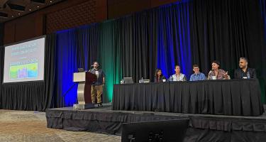 Photo of panelists and organizers listen to Eden Fineday speak during the plenary