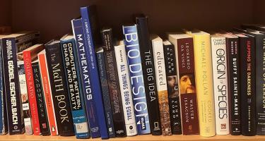 Rectangular photo of Patchen Barss’ office bookshelf with titles on randomness, mathematics, computers, patterns, chaos, and biodesign. The shelf includes NASW author Kenneth Miller’s book Mapping the Darkness. Photo credit: Patchen Barss.