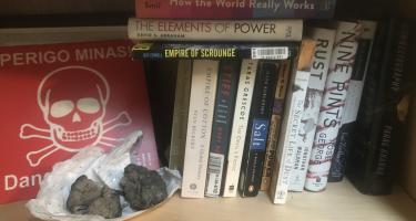 Rectangular photo of Vince Beiser’s office bookshelf showing books on mining cotton, salt, and metals, along with three polymetallic nodules, that is, rocks containing cobalt, nickel, and copper from the Pacific Ocean floor. The shelf also contains a warning sign from a minefield in Angola. Photo credit: Vince Beiser.