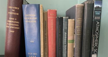 Rectangular photo of Carl Zimmer’s office bookshelf showing works on Anthrax, Black Death, airborne infections, airborne contagion, disease maps, biohazards, and atmospheric science. Photo credit: Carl Zimmer.