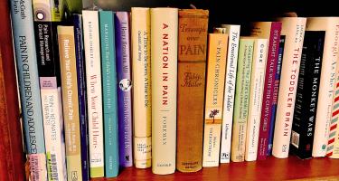 Rectangular photo of a row of books on a wooden bookshelf, with many titles related to the science of pain and children's health, some new and some visibly old.