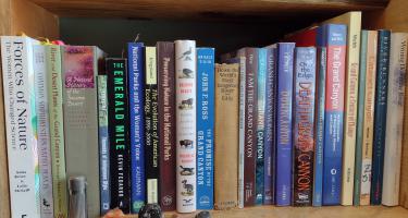 Rectangular photo of Melissa Sevigny’s office bookshelf showing books on the Grand Canyon, botany, and women in science, with waterproof silver match case used by Lois Jotter on 1938 expedition and four Arizona rocks. Photo credit: Melissa Sevigny.