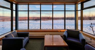 Horizontal photo of an empty atrium, with mid-century modern furniture looking out through expansive, wide windows into a snowy winter field.