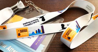 Horizontal photo of a Science Writers 2022 name badge and lanyard draped artistically on a wood-grain table top. The conference logo and St. Jude logo are visible, along with an attendee ribbon attached to the name badge.