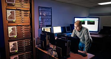 Photograph of a man working in a dimly lit office full of computer server banks and screens.