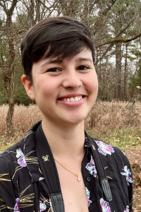 Portrait photo of Ariana Remmel smiling and wearing a floral shirt standing out in a pine and grassland forest