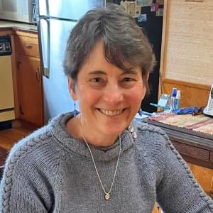 Square photo of Jennifer Huber smiling and wearing a sweater seated in a kitchen.