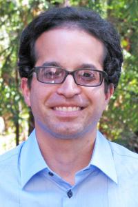 Portrait photo of Ramin Skibba wearing glasses, collared shirt, and posing outdoors in greenery